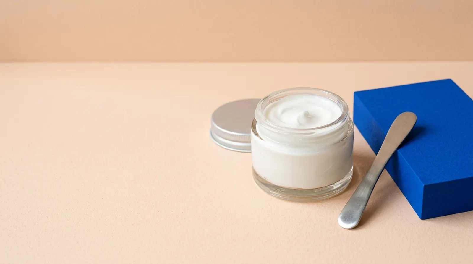 Unbranded cream jar, stainless spatula, and cobalt paper on a pale peach work surface